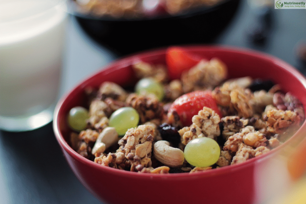A close-up image of a red bowl filled with granola, green grapes, cashews, strawberries, and dried fruits, with a glass of milk and another bowl in the blurred background.