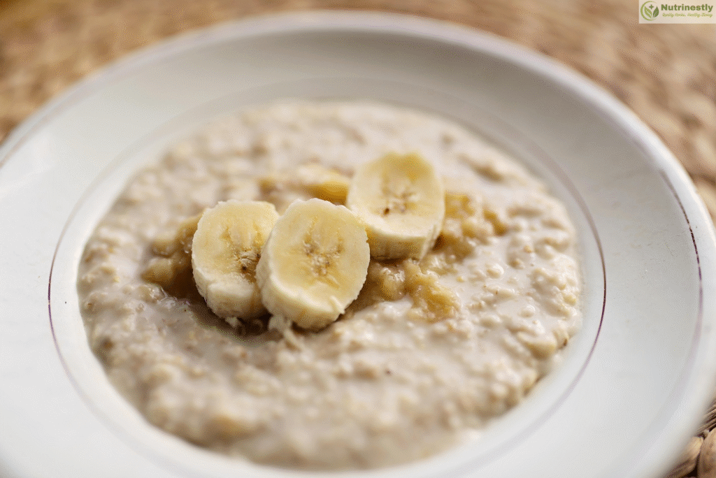 A close-up image of a bowl of creamy oatmeal topped with three slices of banana, served in a white dish with a faint red rim. rich in fiber .
