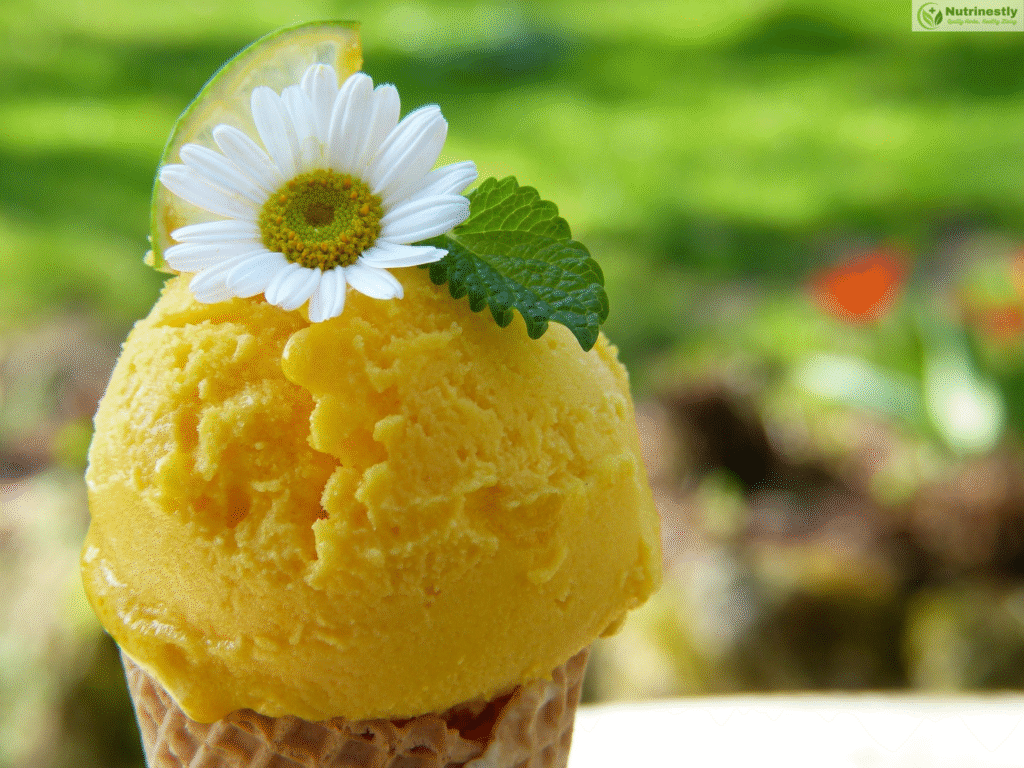 "Close-up of a scoop of Mango ice cream in a waffle cone, garnished with a daisy flower, mint leaf, and lime slice, with a blurred garden background.