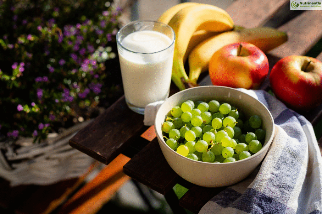 A bowl of fresh green grapes, a glass of milk, bananas, and red apples arranged on a wooden outdoor table with a striped cloth, next to a potted plant with purple flowers.