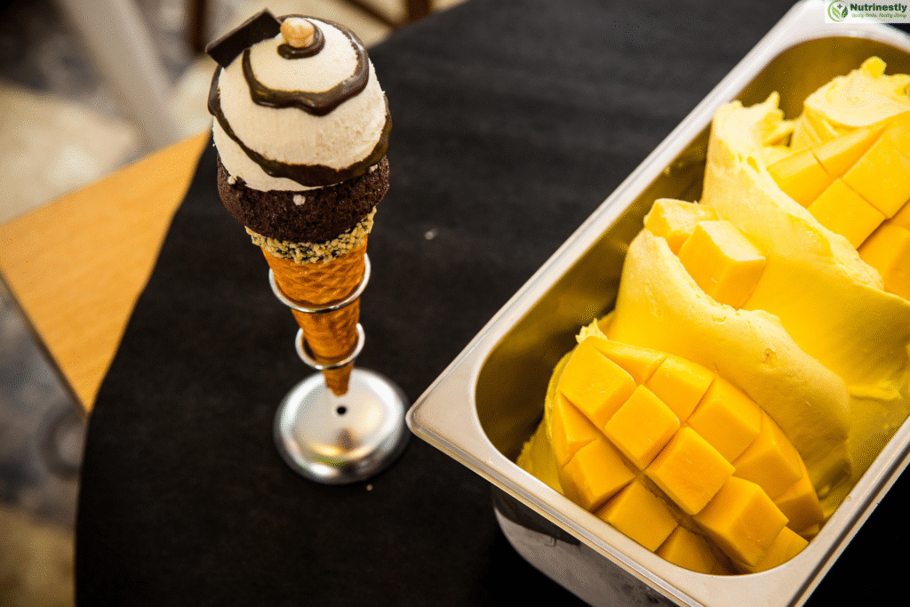 "A serving container filled with creamy Mango ice cream garnished with fresh mango cubes, placed beside a decorative ice cream cone topped with vanilla scoop, chocolate drizzle, and a brownie layer.