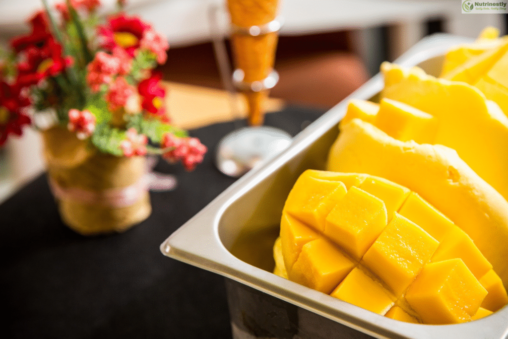 Fresh Mango ice cream served in a metal container, garnished with ripe mango cubes, with a decorative flower vase and an ice cream cone blurred in the background."