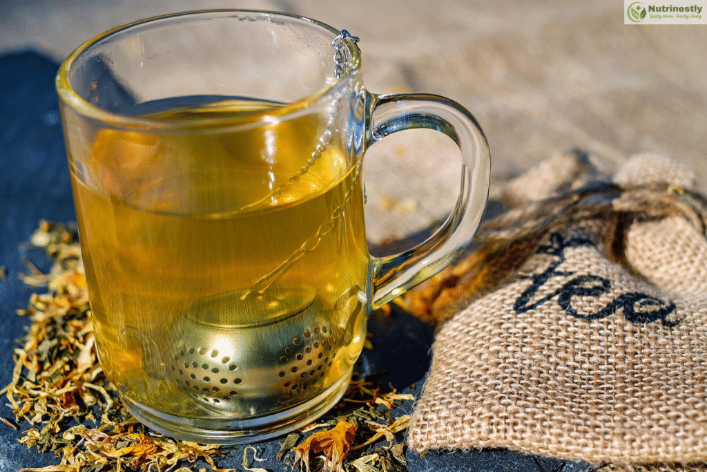 Green Tea or Black Tea : A clear glass mug filled with freshly brewed green tea, with a metal tea infuser inside and loose tea leaves scattered nearby on a rustic surface