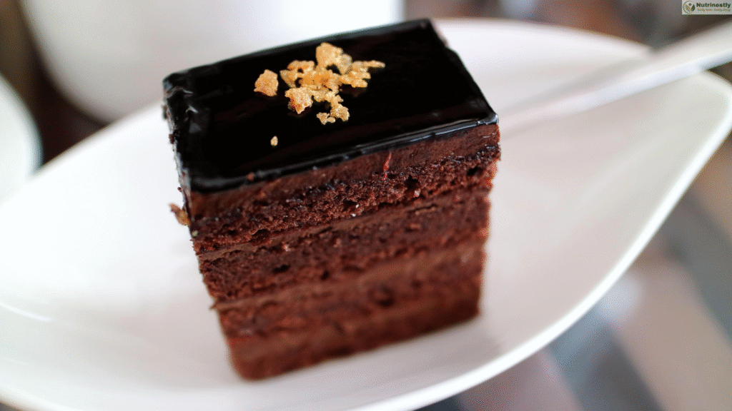 A close-up of a slice of rich, multi-layered, chocolate coffee-flavored cake with glossy dark chocolate ganache on top, garnished with golden flakes, served on a white curved plate.