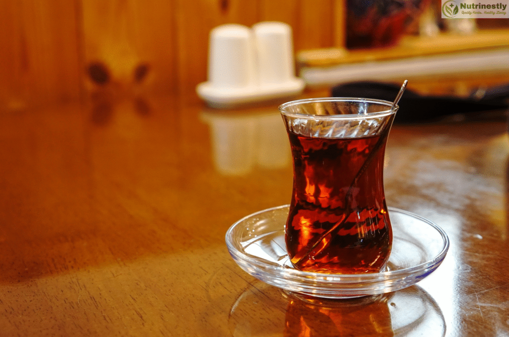 A traditional clear glass of black tea served in a curved glass on a small glass saucer, placed on a wooden table in a cozy setting.
Black Tea vs. Green Tea.