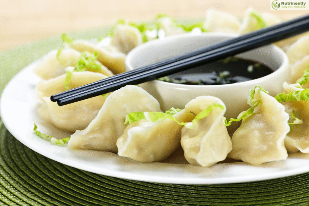 Close-up of steamed Chinese dumplings arranged on a white plate, garnished with shredded lettuce and served with soy dipping sauce and chopsticks.