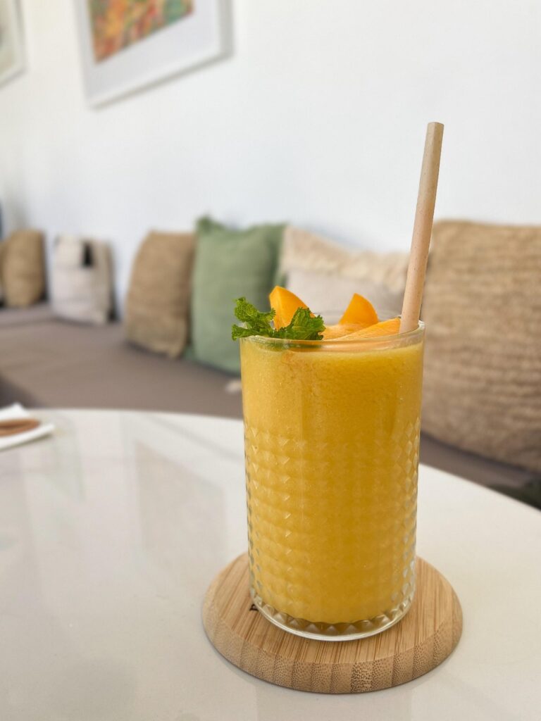 Chilled mango smoothie served in a textured glass with mint leaves and mango slices as garnish, placed on a wooden coaster in a cozy cafe setting.
