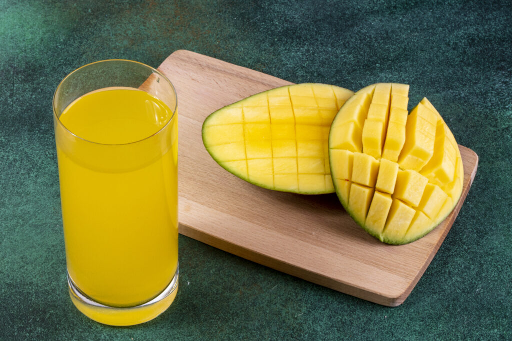 A glass of fresh mango juice next to sliced ripe mango halves on a wooden board